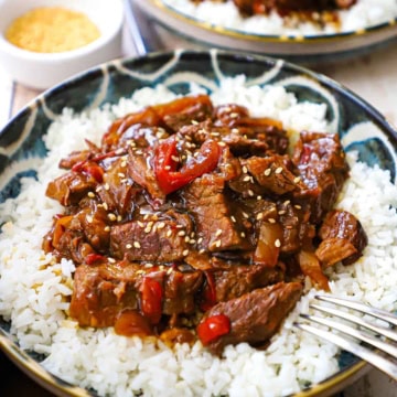 A close-up view of slow cooker pepper steak on top of steamed white rice on colorful shallow bowl.