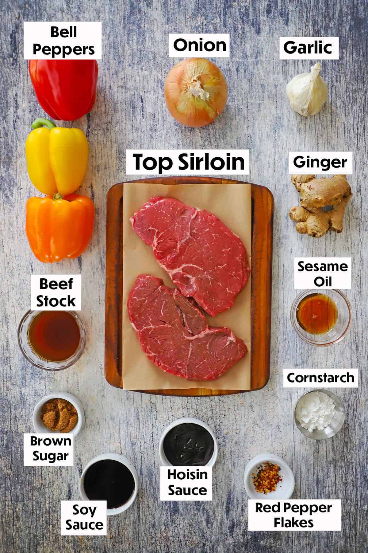 An arrangement of ingredients for slow cooker pepper steak on a grey wooden background including bell peppers, onion, garlic, sirloin, ginger, beef stock, oil, soy sauce, hoisin sauce, and brown sugar.