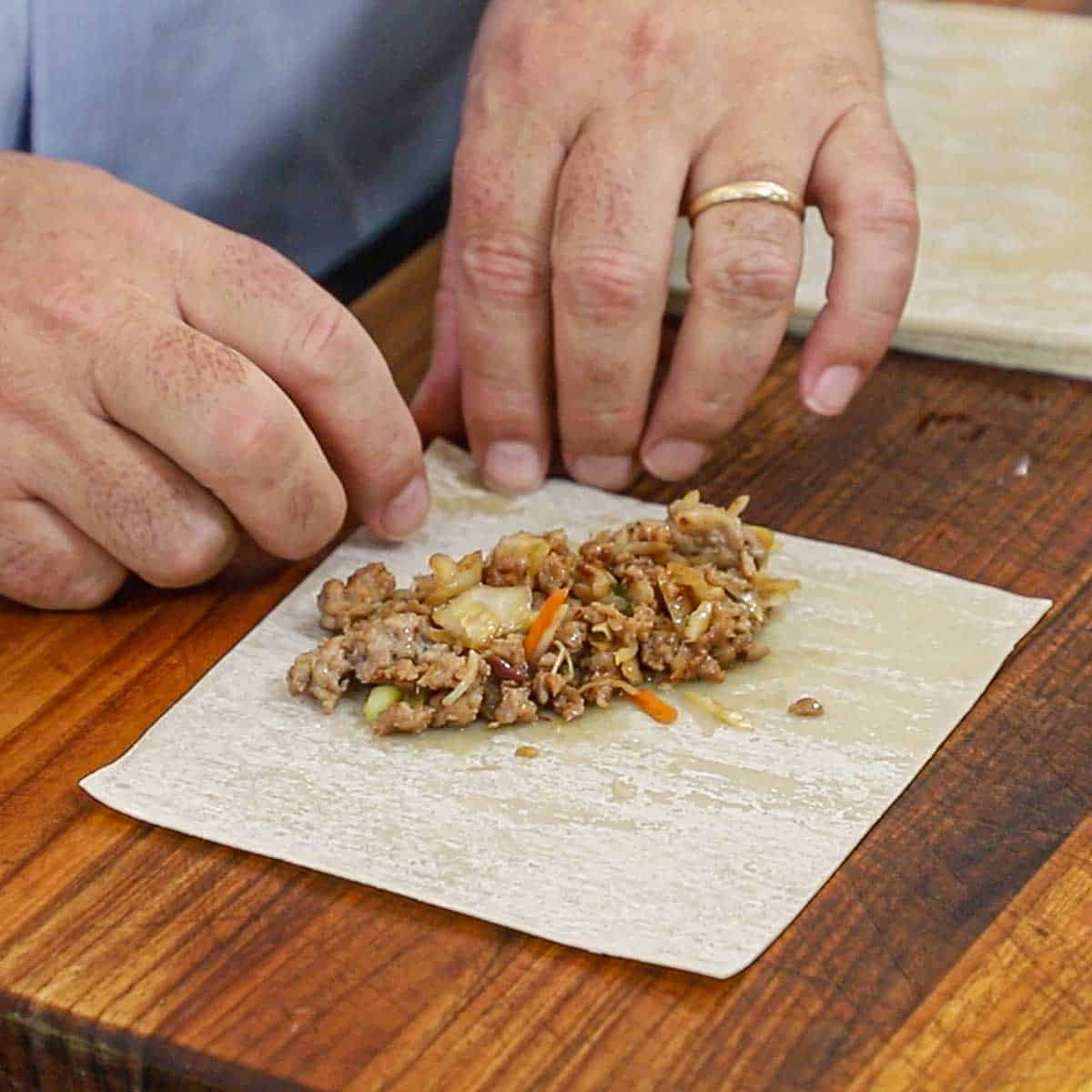 A person getting ready to use fingers to roll the edge of an egg roll wrapper over a pork and cabbage filling the is in the center of the wrapper.