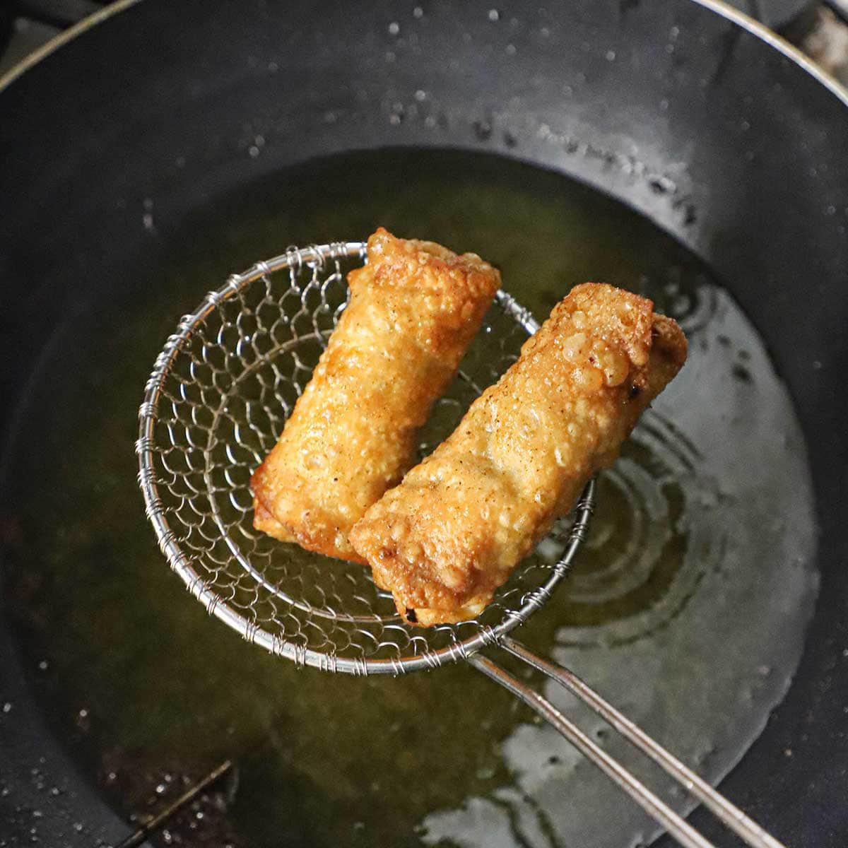 Two crispy freshly made egg rolls being held in a mesh spider over a wok that is filled with hot vegetable oil.