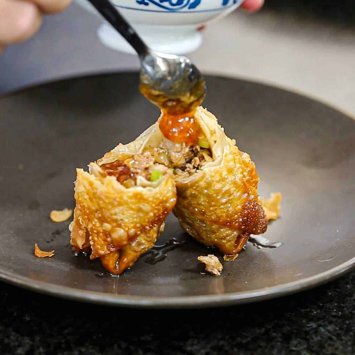 A person using a small spoon to add homemade duck sauce over the top of a homemade egg roll that has been split in half and is sitting upright in the middle of a black dinner plate.