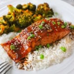A close-up view of a baked teriyaki salmon fillet on a bed of white rice with a side of roasted broccoli next to it all on a white dinner plate.
