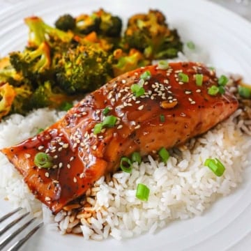 A close-up view of a baked teriyaki salmon fillet on a bed of white rice with a side of roasted broccoli next to it all on a white dinner plate.