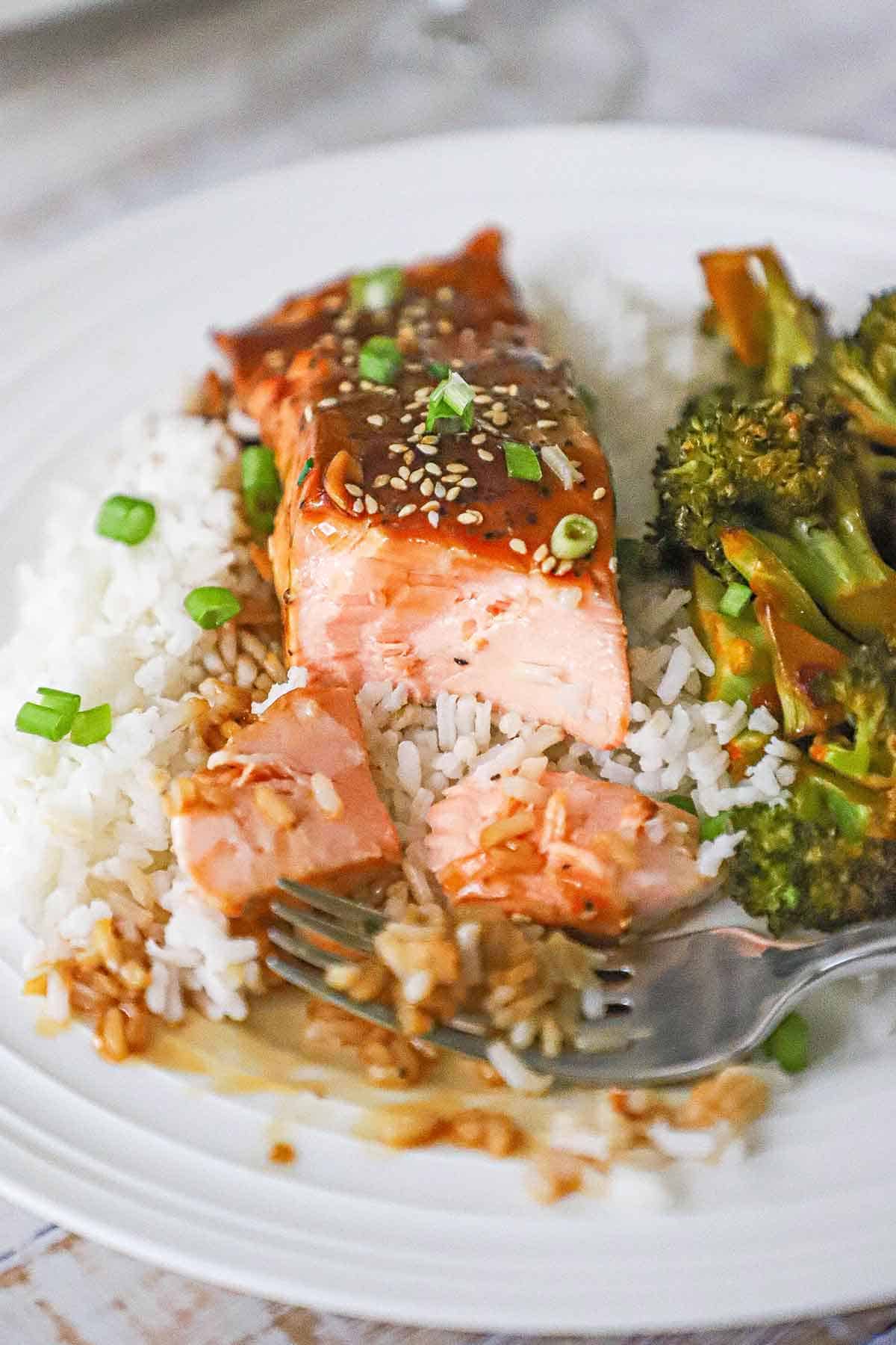 A partially eaten fillet of baked teriyaki salmon resting on a bed of steamed white rice next to roasted broccoli on a white dinner plate.