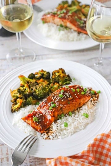 An elegant white dinner plate filled with a serving of baked teriyaki salmon over steamed white rice next to a serving of roasted broccoli with a glass of white wine nearby.