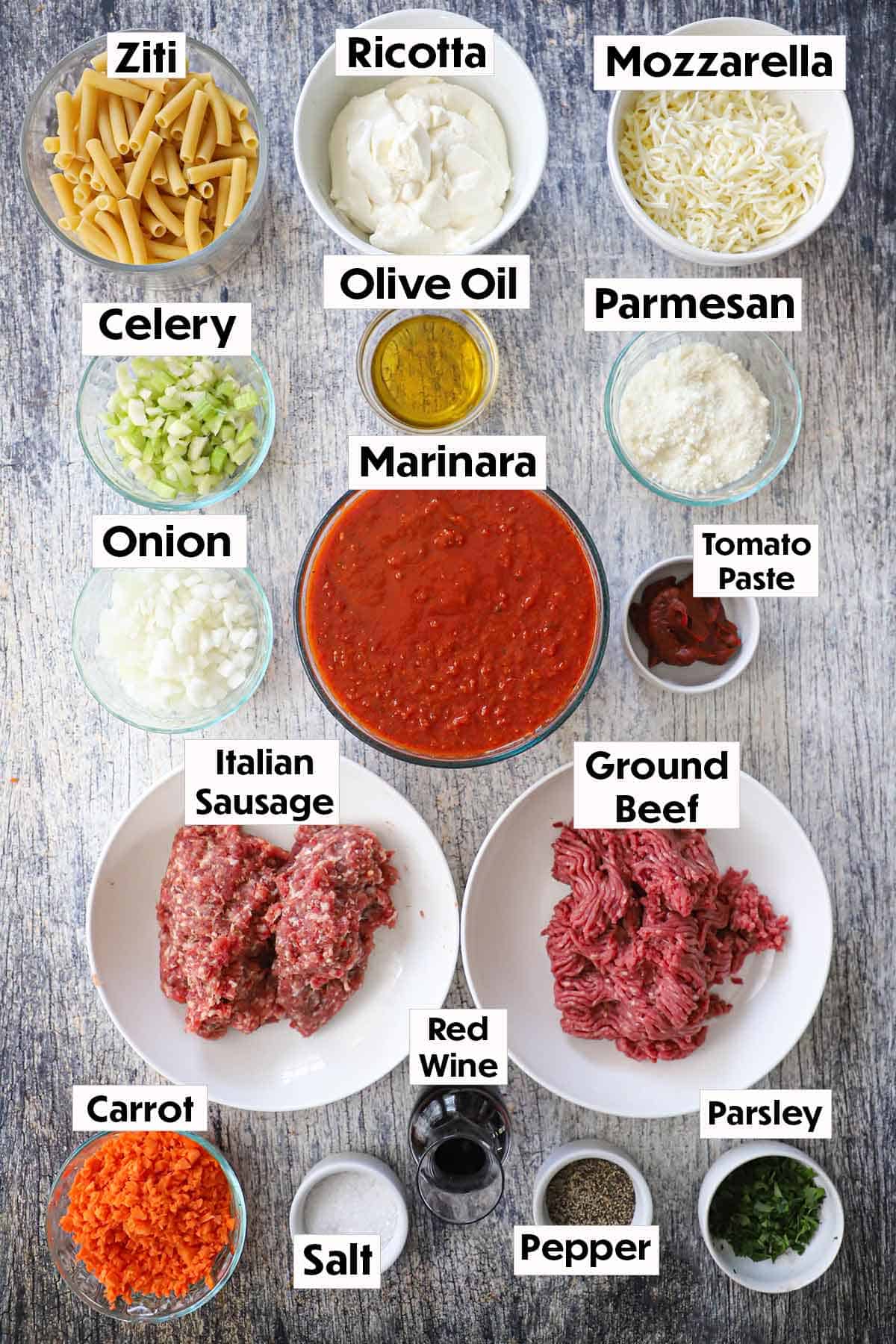 An arrangement of ingredients for best-ever baked ziti on a wooden grey background including uncooked zit, ricotta, mozzarella, celery, onion, oil, marinara sauce, sausage, ground beef, carrots, red wine, and seasonings.