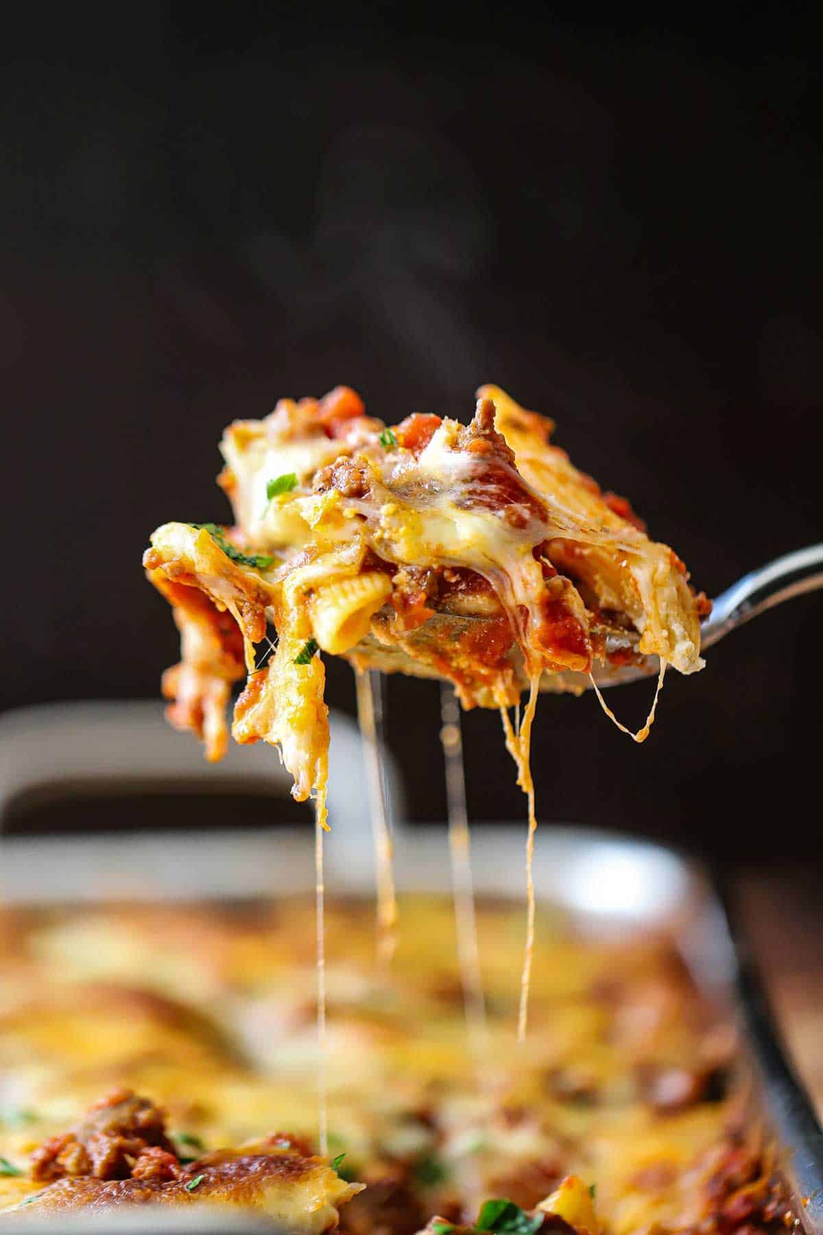 A person raising a cheesy serving of best-ever baked ziti from a pan of the same thing.