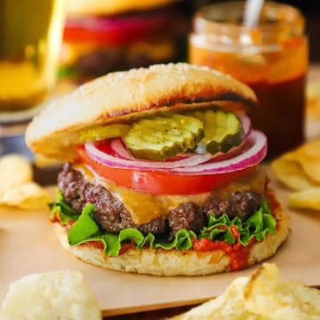 A best-ever cheeseburger topped with a slice of tomato, thin slices of red onion, bread and butter pickles, resting on green leaf lettuce, homemade ketchup, all inside a homemade hamburger bun.