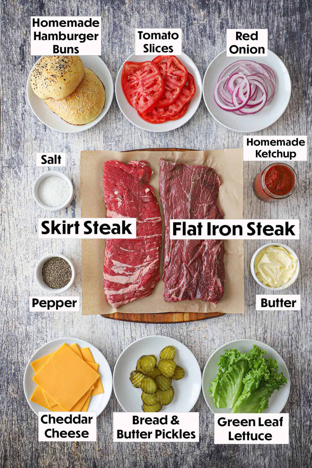 An arrangement of ingredients on a grey wooden background for best-ever cheeseburger including skirt steak, flat-iron steak, buns, tomatoes, onion, cheese, pickles, seasonings, and homemade ketchup. 