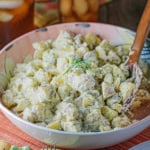 A colorful serving bowl filled with homemade best-ever potato salad with fresh dill and a wooden spoon inserted into the side of the salad.
