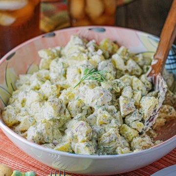 A colorful serving bowl filled with homemade best-ever potato salad with fresh dill and a wooden spoon inserted into the side of the salad.