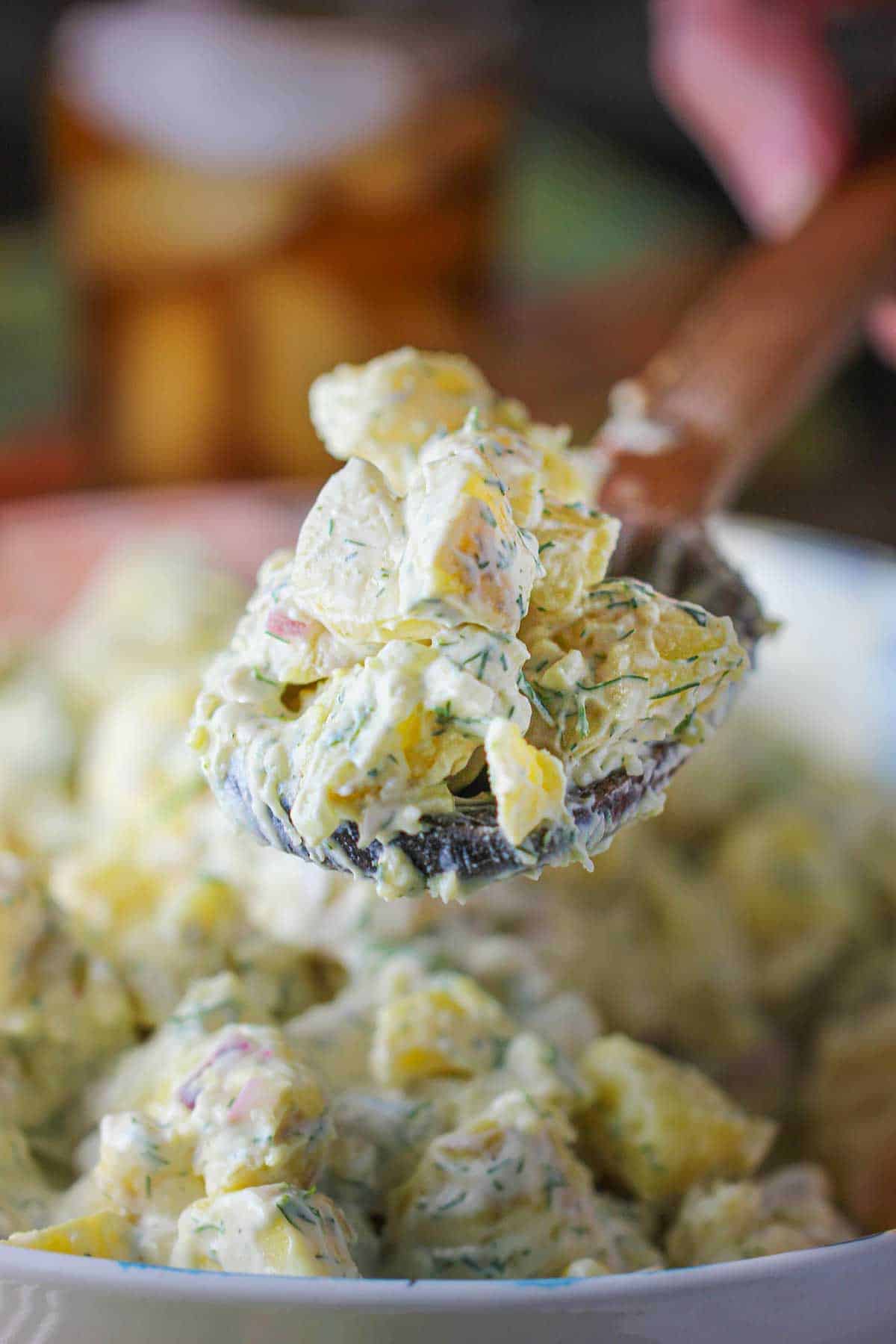 A person raising a wooden spoon filled with a serving of best-ever potato salad over a bowl of the same.