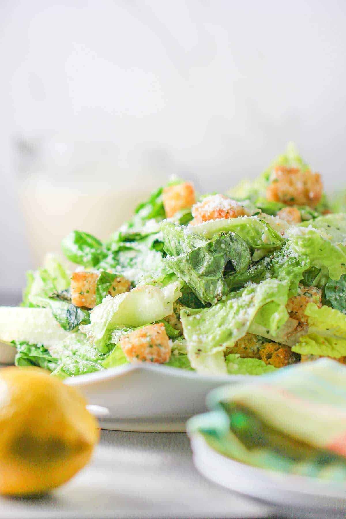 A large classic Caesar salad sitting on a white platter with a whole lemon and dinner plates nearby.