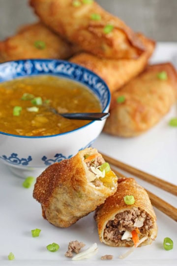 A homemade egg roll that has been split in half revealing the pork and cabbage inside with one of the halves resting on the other half with a bowl of homemade duck sauce in the background.