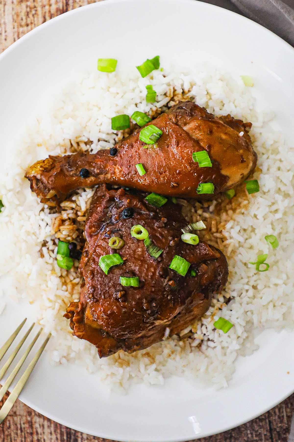 A serving of Filipino chicken adobo including a chicken leg and thigh resting on a bed of steamed white rice and garnished with chopped scallions all on a white dinner plate.