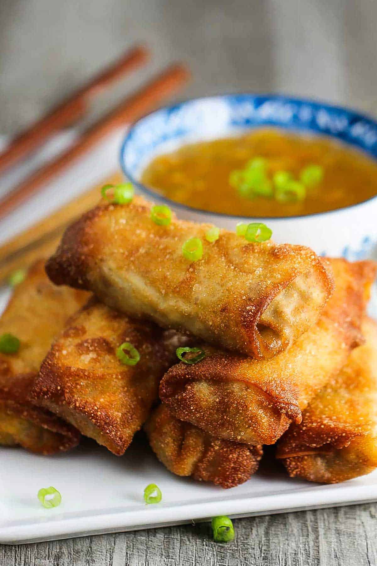A pile of homemade egg rolls on a white platter with chopped scallions sprinkled over the top and a bowl of homemade duck sauce in the background.