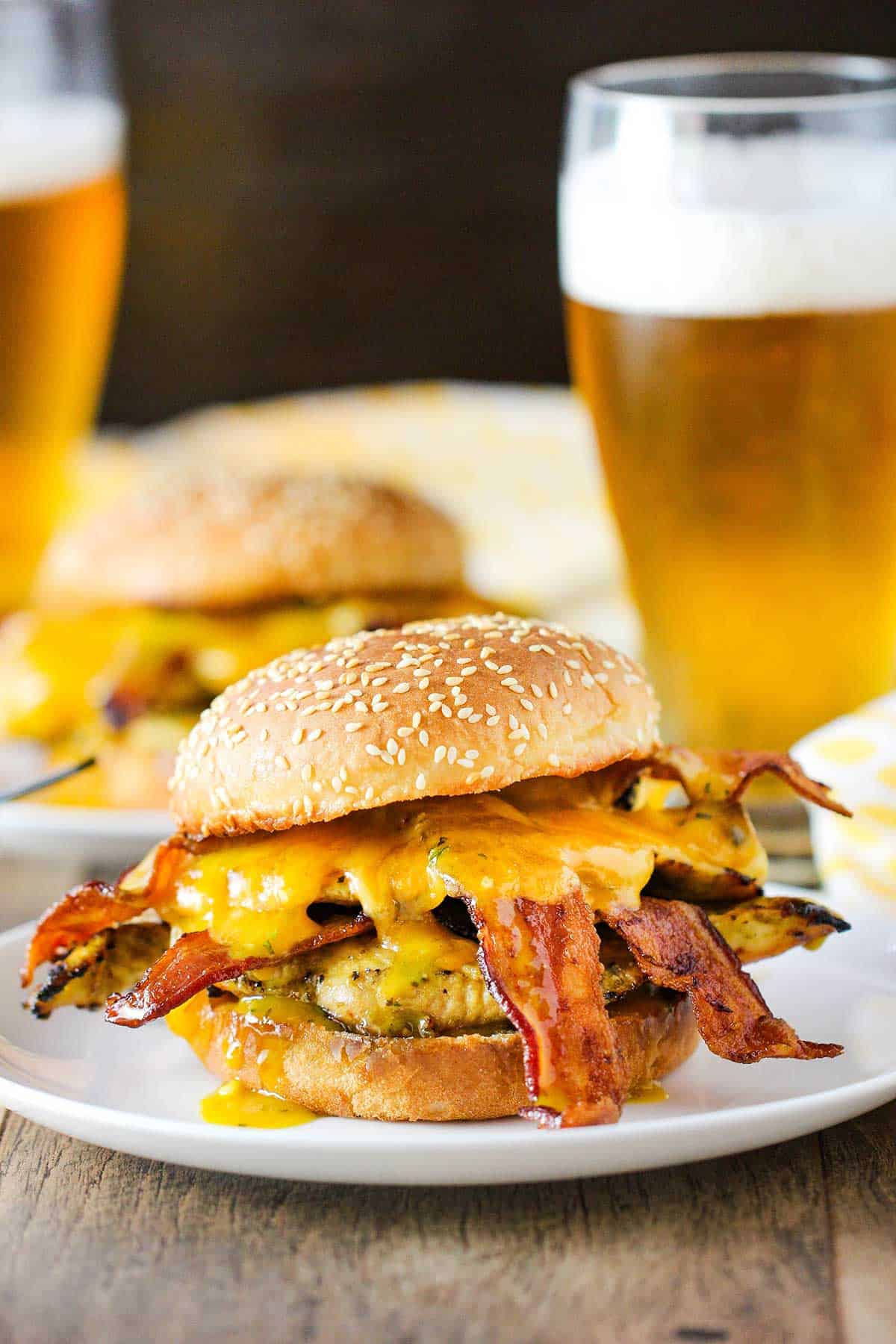 A grilled honey mustard chicken sandwich with bacon on a sesame seed bun sitting on a withe dinner plate with a frosty glass of beer in the background.