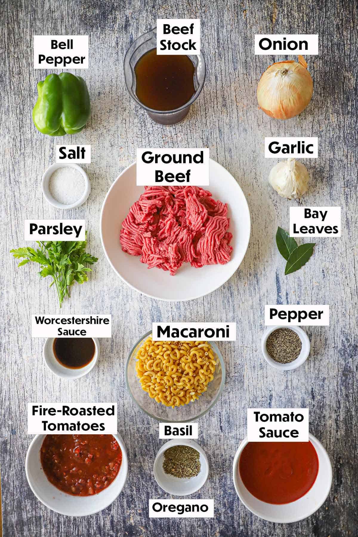 An arrangement of ingredients for Mom's goulash (American-style) on a wooden grey background including ground beef, macaroni pasta, bell pepper, onion, garlic, beef stock, tomato sauce, diced tomatoes, and herbs.