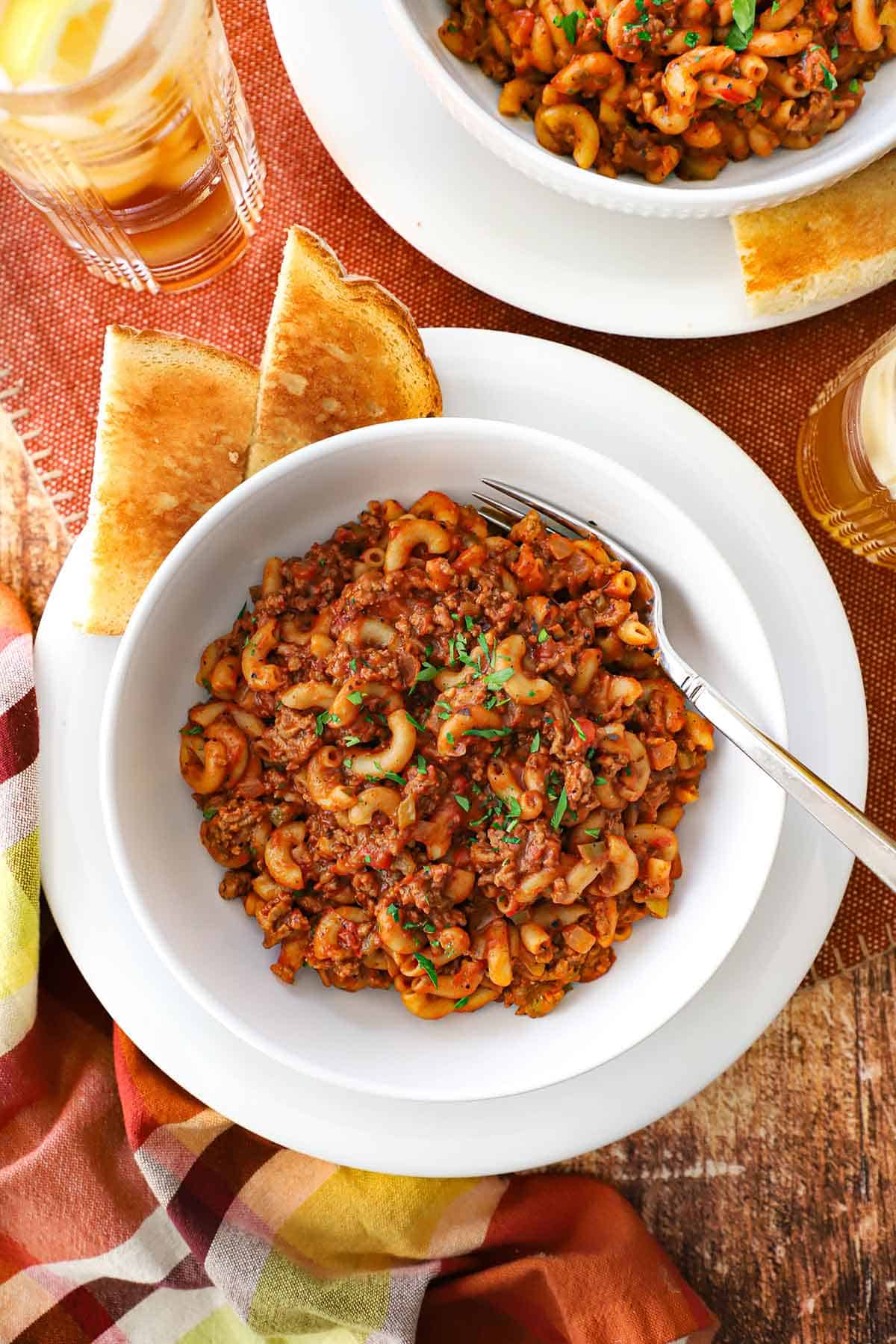 A white bowl filled with Mom's goulash (American-style) sitting on a white dinner plate with two pieces of toast resting on the plate next to the bowl.