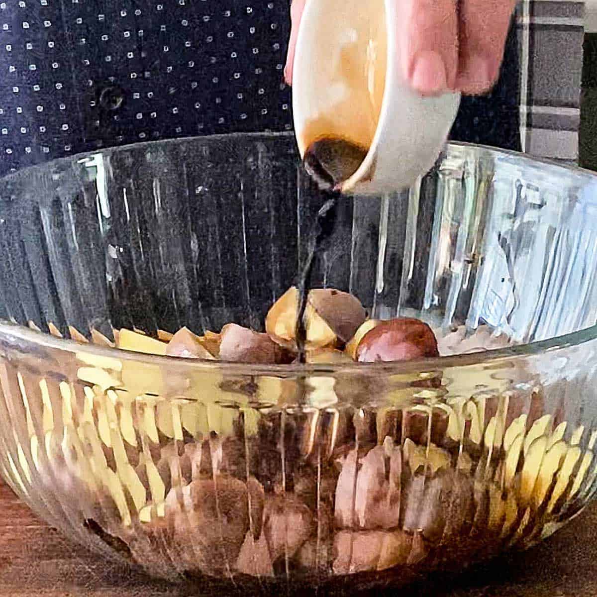 A person transferring balsamic vinegar from a small white bowl into a large glass bowl filled with new potatoes that have been cut in half.