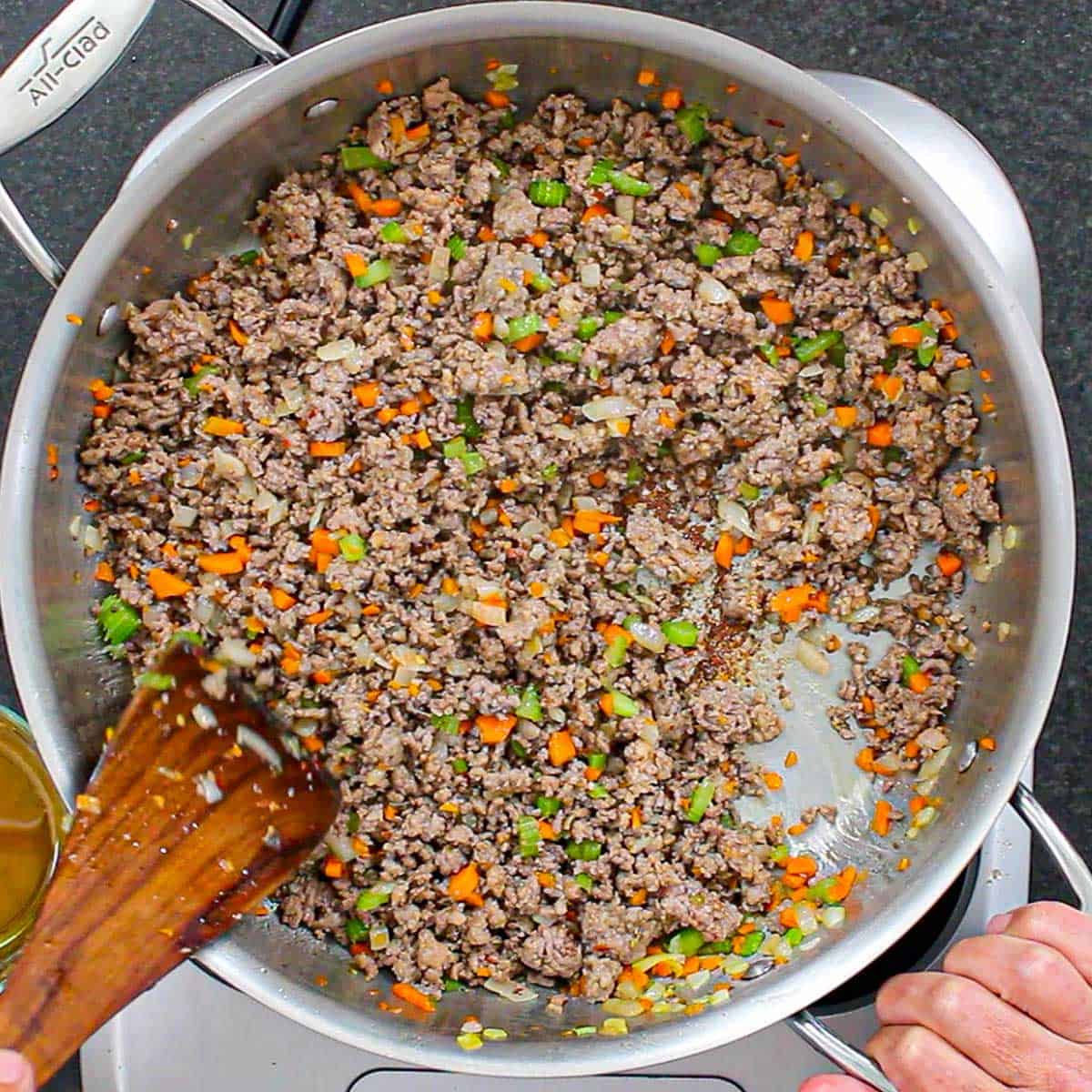 A person stirring cooked crumbled Italian sausage, ground beef with chopped carrots, celery, and onion with a wooden spatula in a large stainless steel saucepan.