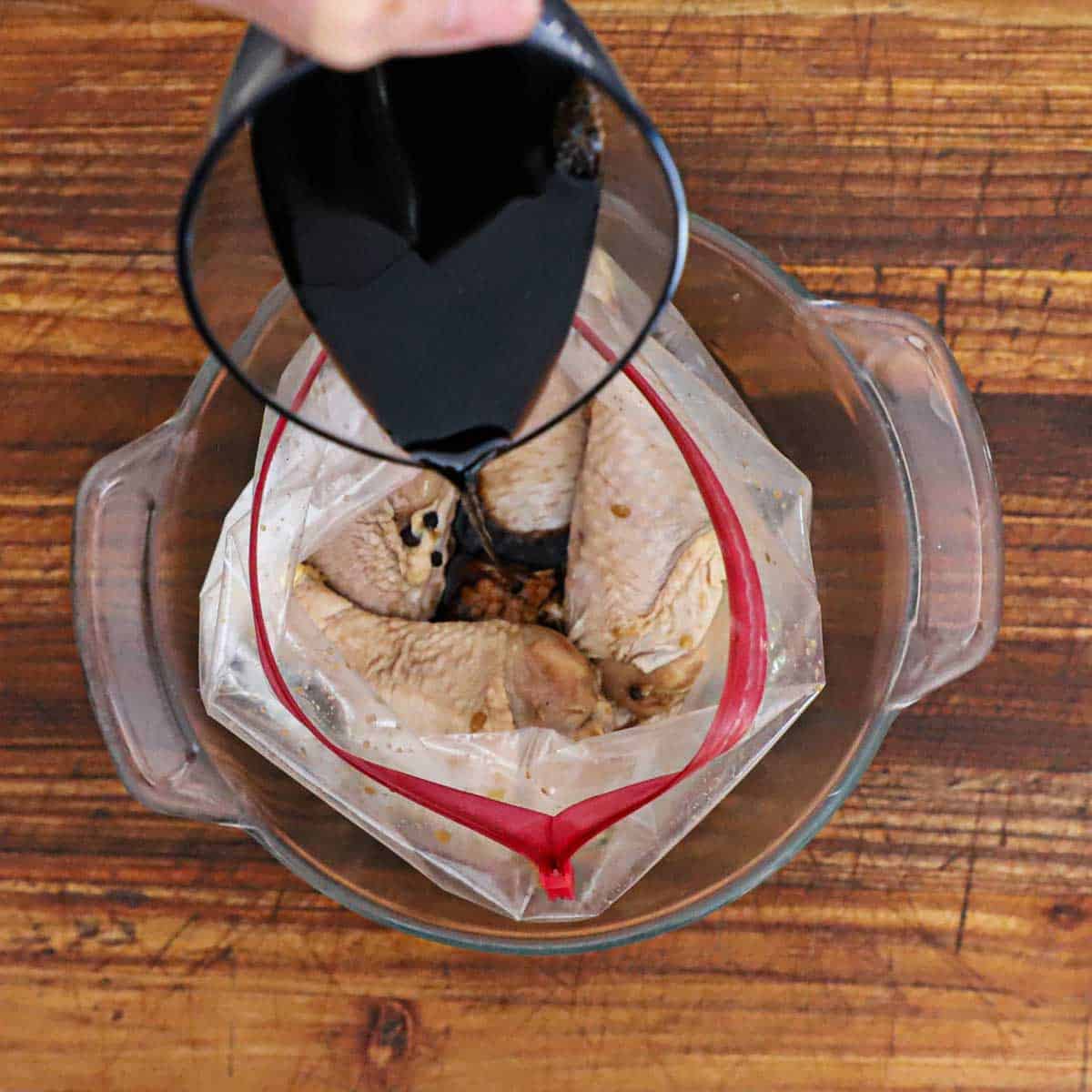 A person pouring soy sauce into a large freezer baggie that is filled with marinated chicken pieces.