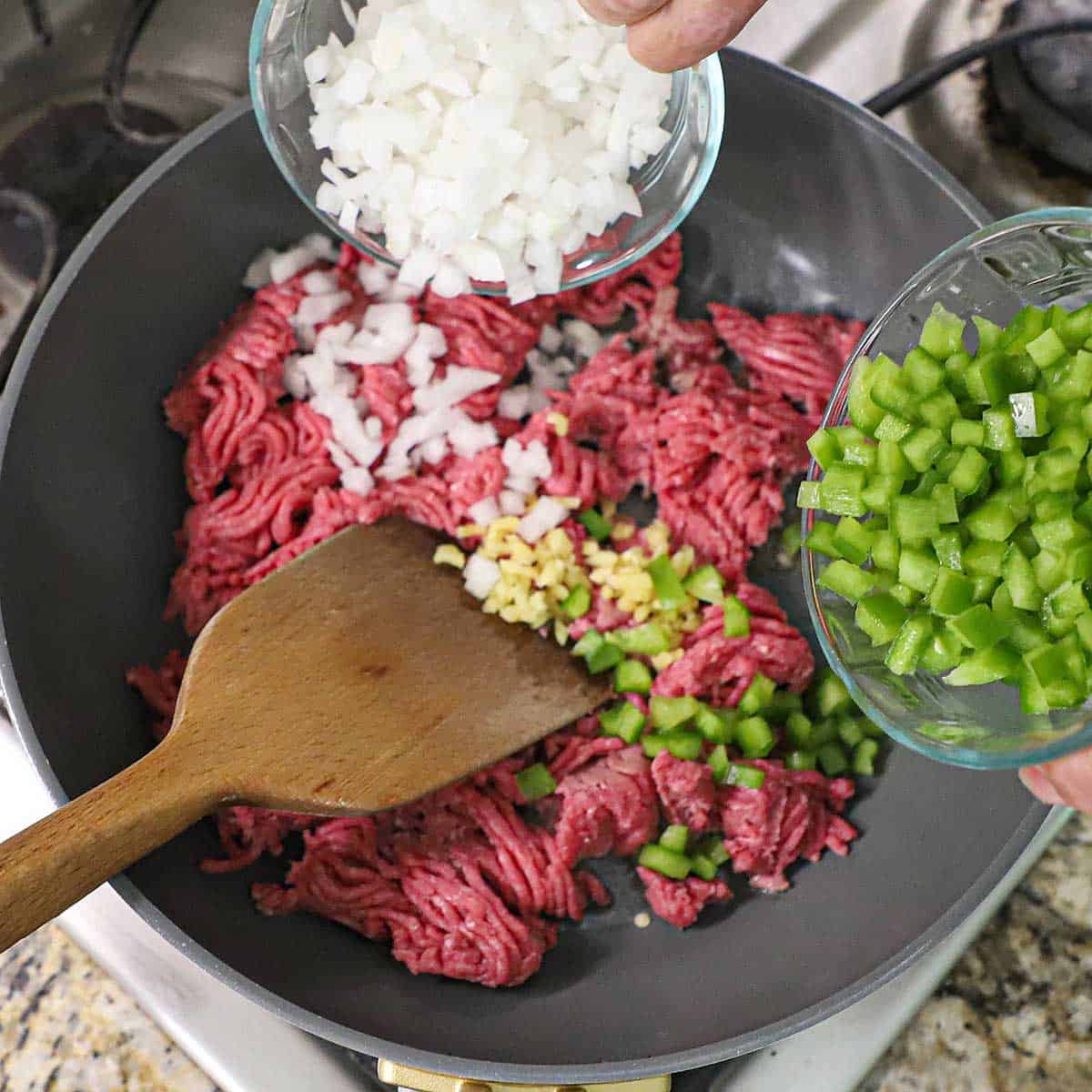 A person adding chopped onion from one glass bowl and chopped green bell pepper from another glass bowl into a skillet that had ground beef and minced garlic in it.