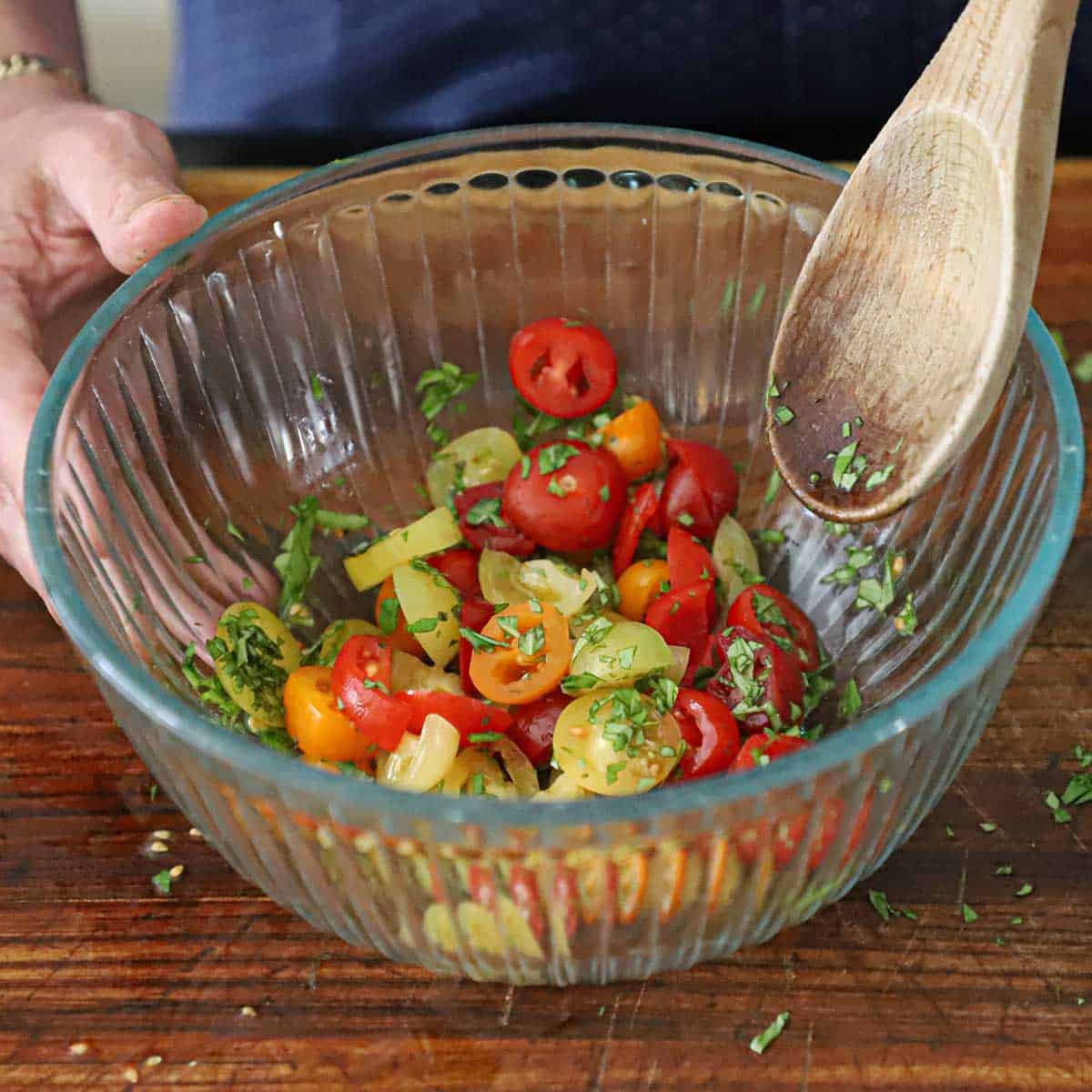 A person using a wooden spoon to mix halved red, yellow, and orange cherry tomatoes with chopped fresh basil.