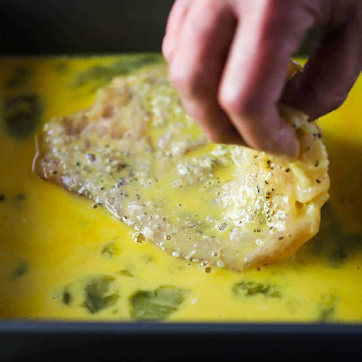 A person using his fingers to soak a thin chicken cutlet that had been coated with flour in an egg bath in a metal pan.