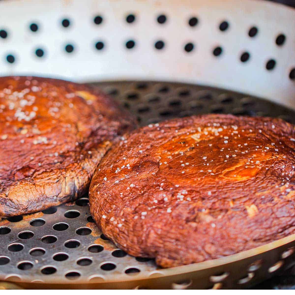 Two large portobello mushroom caps being grilled in a grill pan on a gas grill.