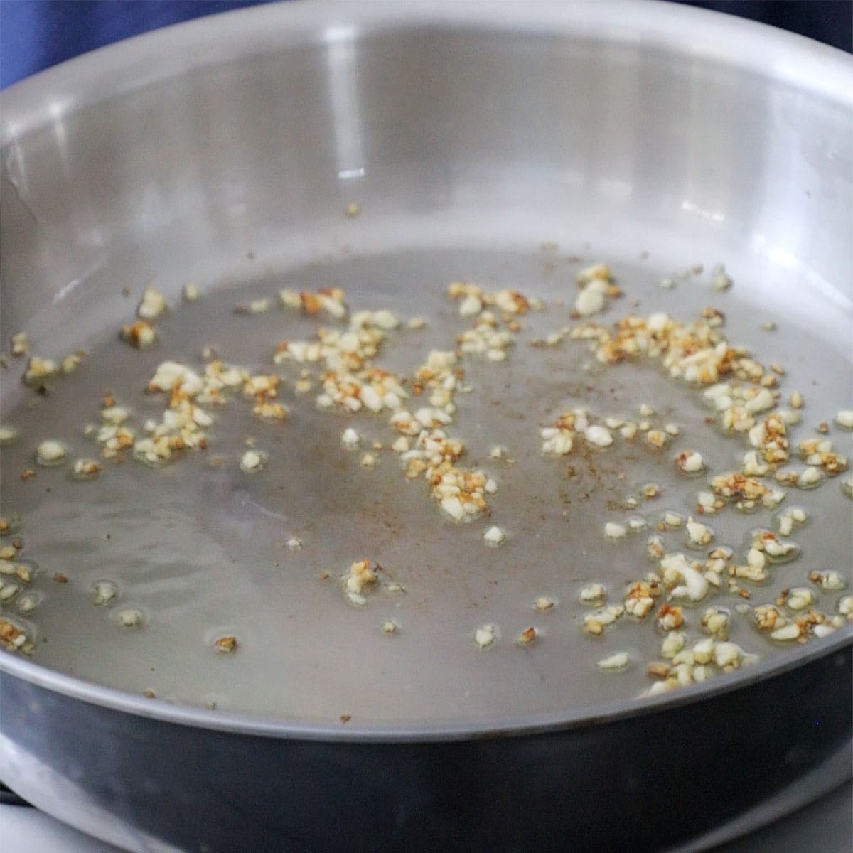A large stainless steel skillet with minced garlic simmering in olive oil on a gas stove.