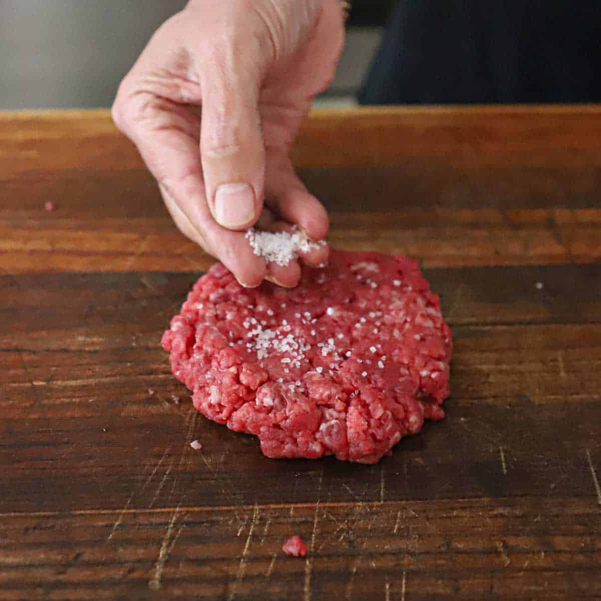 A person sprinkling coarse Kosher salt over the top of a freshly formed hamburger patty resting on a wooden cutting board.