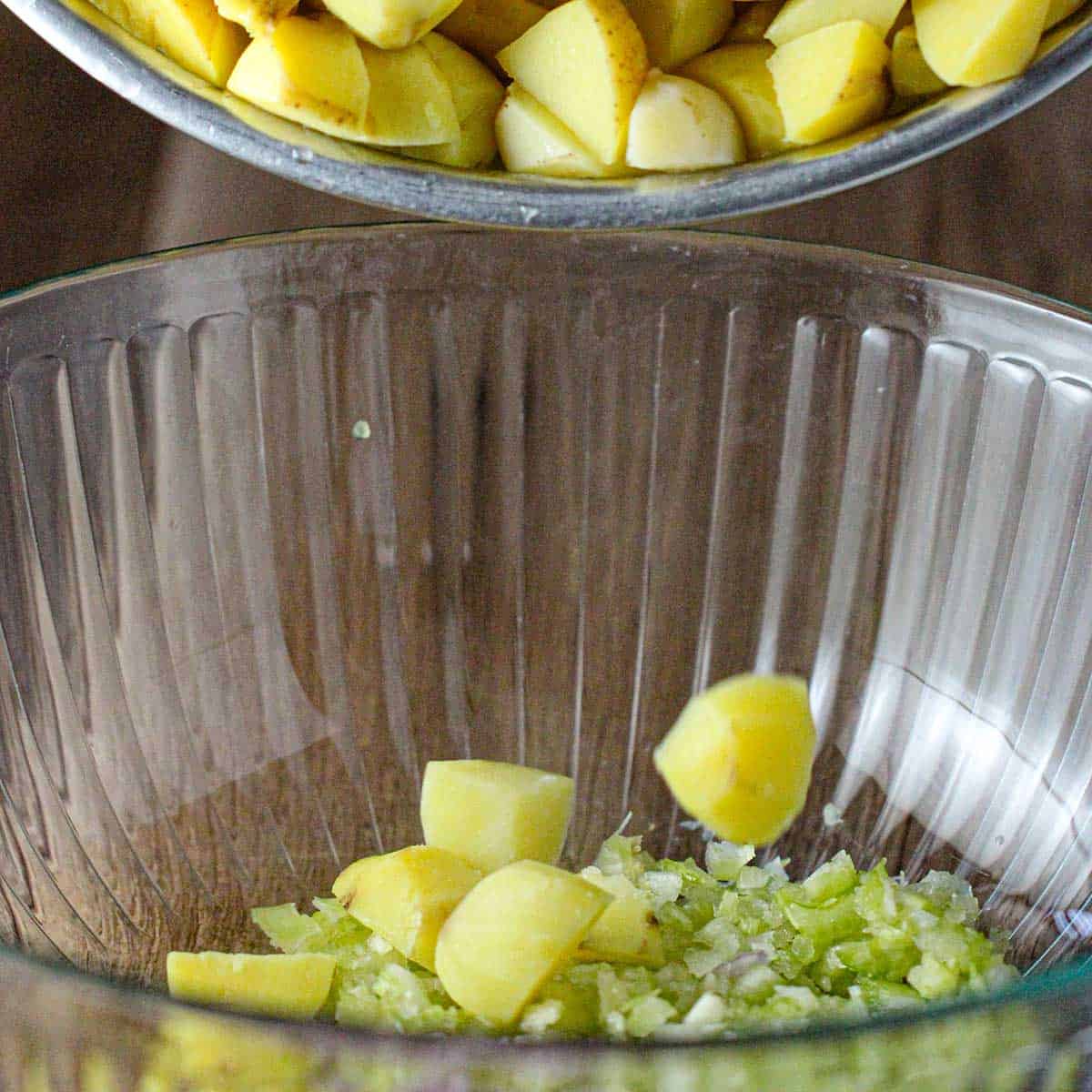 A person dumping freshly boiled cubes of yellow potatoes into a glass bowl with chopped onion and celery in the bottom.