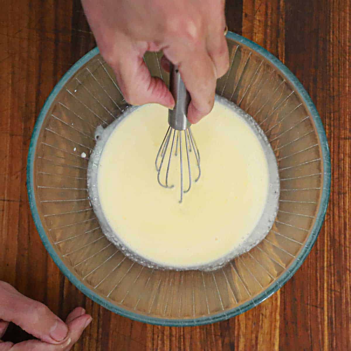 A person using a whisking egg yolks with heavy cream in a glass bowl on a wooden cutting board.