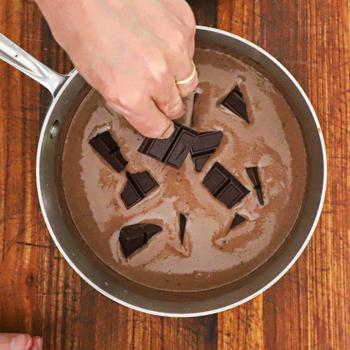 A person adding pieces of semi-sweet chocolate into a saucepan that is filled with a warm chocolate cream sauce.