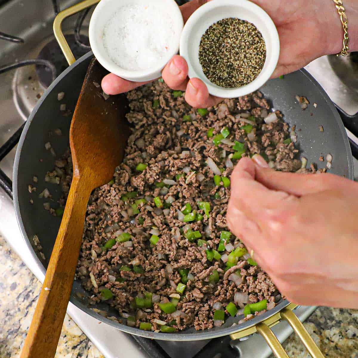 A person holding small bowls of salt and pepper over a skillet filled with browned ground beef with sautéed bell peppers and onions in it.