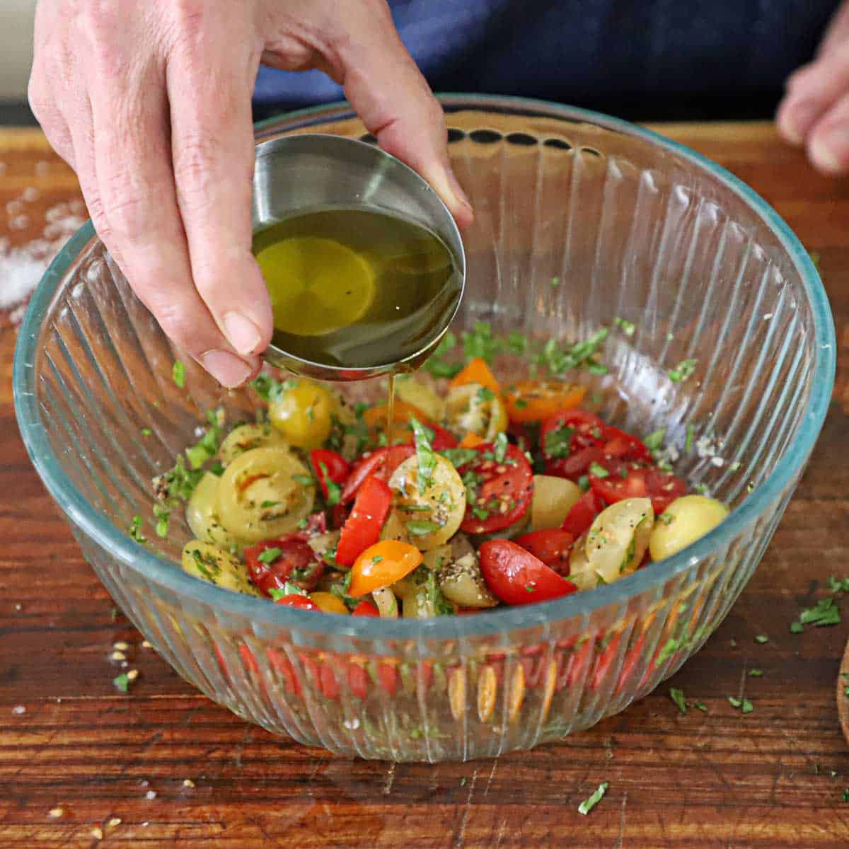 A person pouring extra-virgin olive oil into a glass bowl that is filled with a mixture of halved cherry tomatoes, chopped fresh basil, salt, and pepper.
