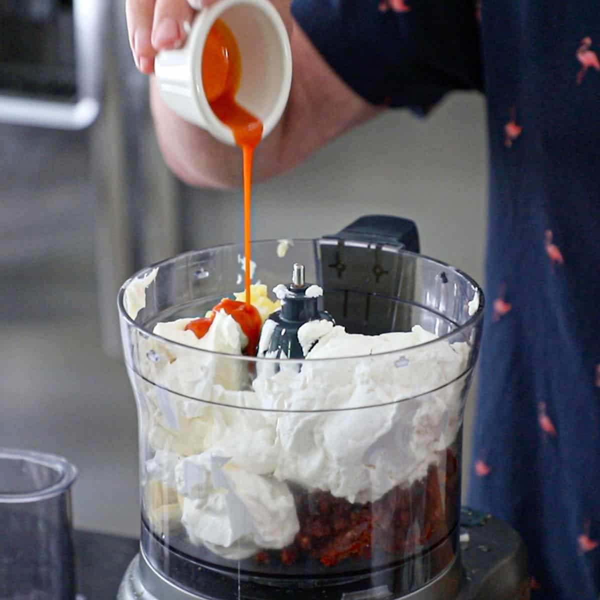 A person pouring a small amount of hot sauce from a small white bowl into a food processor containing softened cream cheese, goat cheese, sour cream, garlic, artichoke hearts, and sun-dried tomatoes.