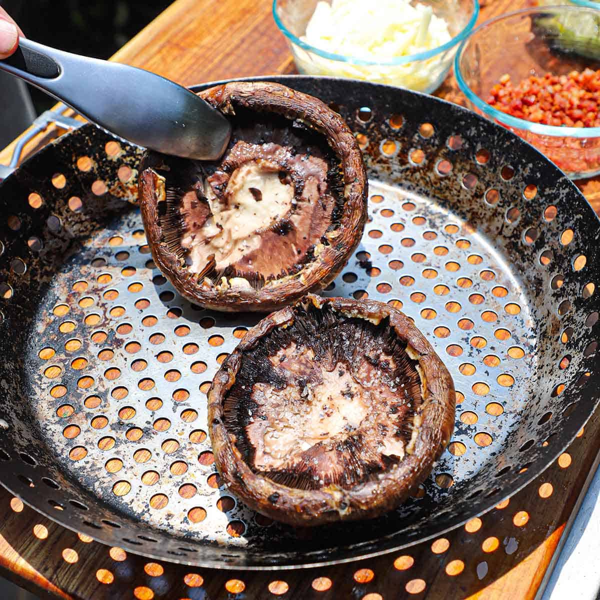 A person using a pair of tongs to flip a couple of grilled portobello mushroom caps that are resting in a grill pan on a wooden cutting board with bowls of pancetta and cheese nearby.
