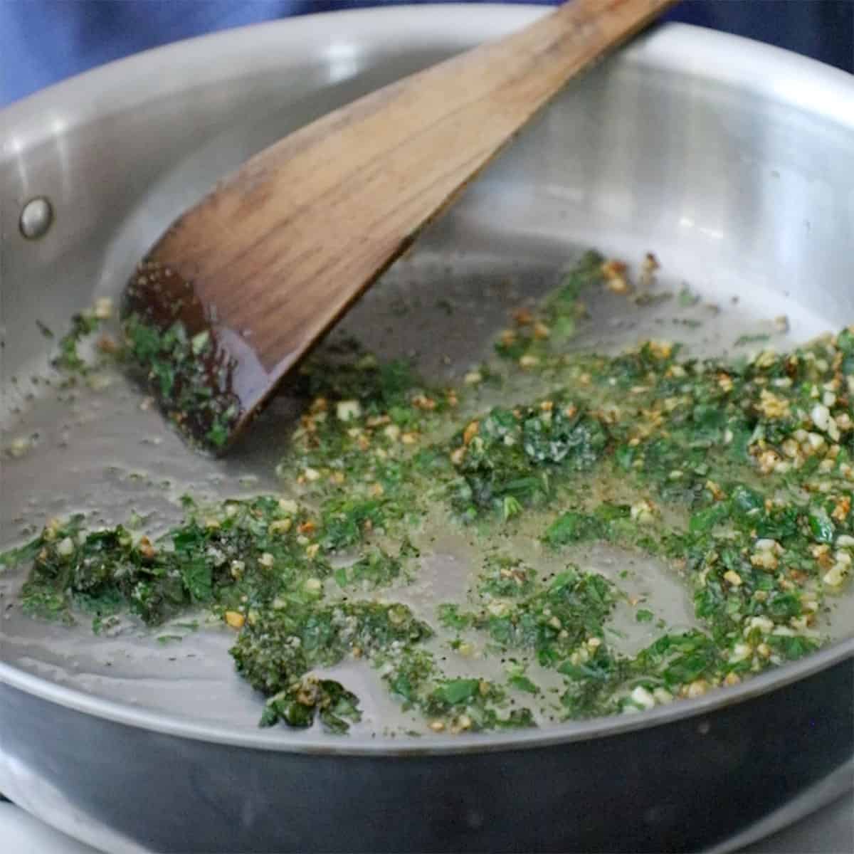 A person using a wooden spatula to stir fresh herbs that are being sautéed with minced garlic that has also been sautéed.