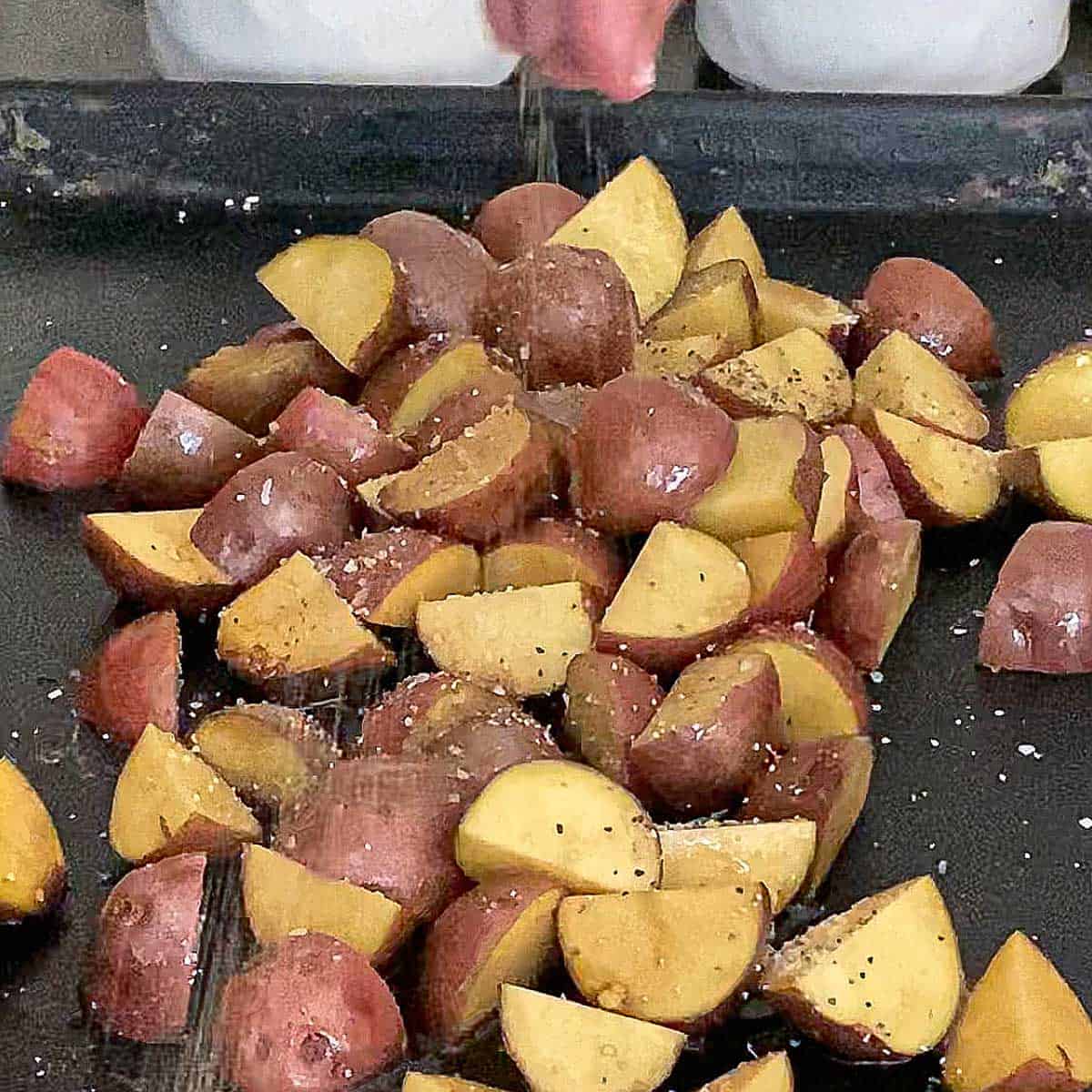 A person sprinkling salt and pepper onto new potatoes that have been halved and are resting on a black baking sheet pan.