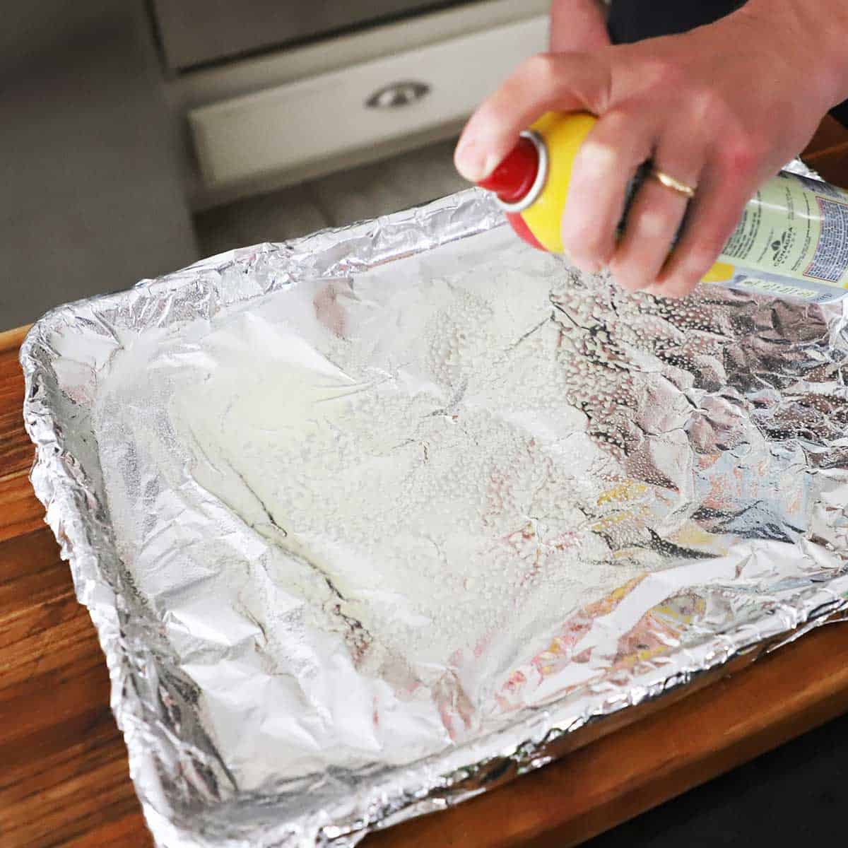 A person spraying a baking sheet that is lined with foil with cooking spray.