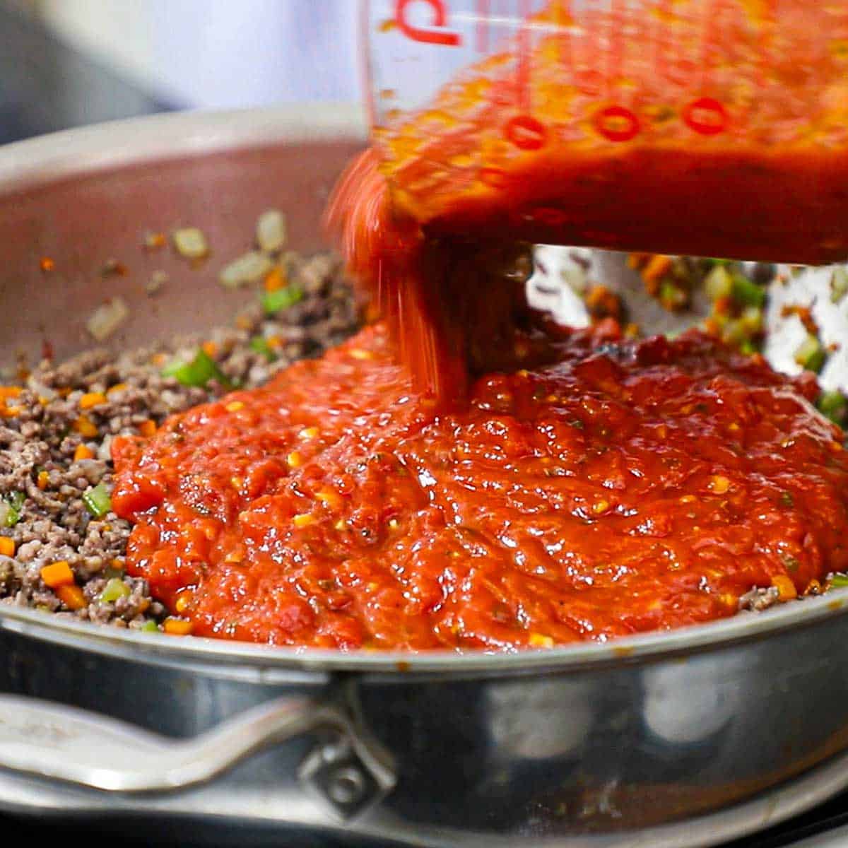 A person pouring a large amount of homemade marinara sauce from a 8-cup measuring cup into a skillet filled with simmering cooked meat and vegetables.