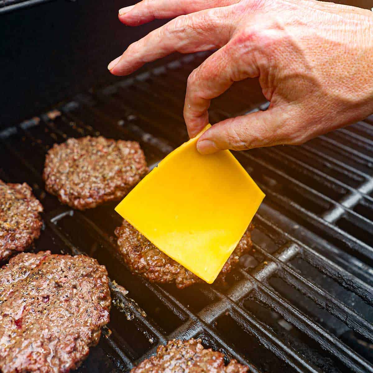 A person placing a slice of cheddar cheese over a grilled hamburger patty that is sitting next to four other grilled hamburger patties.