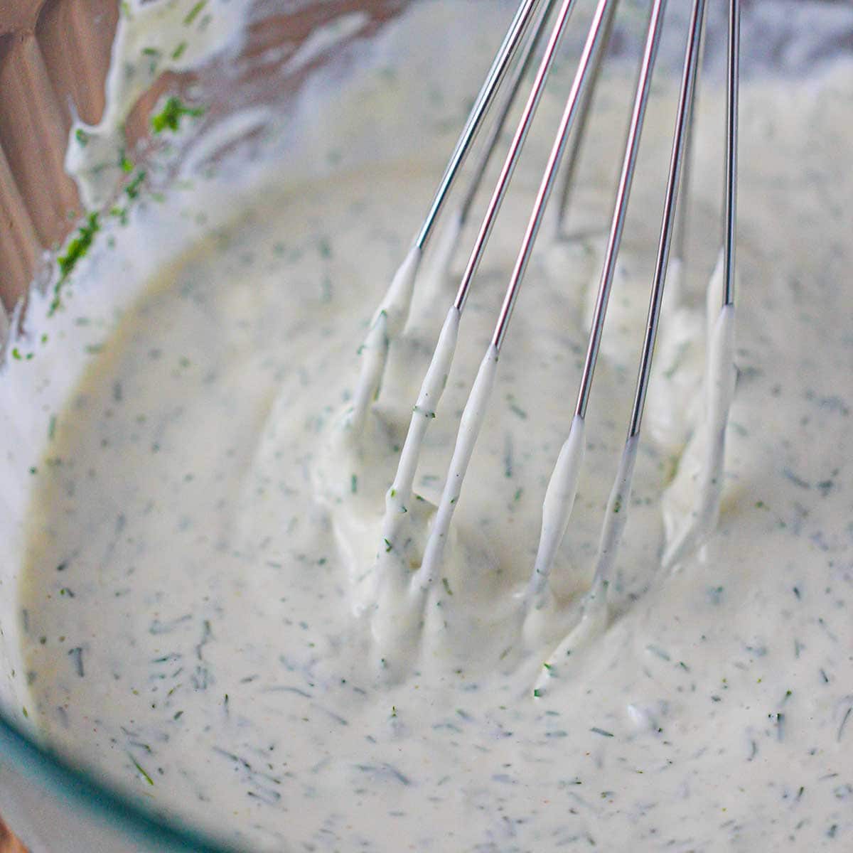 A whisk being used to stir the dill and mayonnaise dressing in a glass bowl that will be used for potato salad.