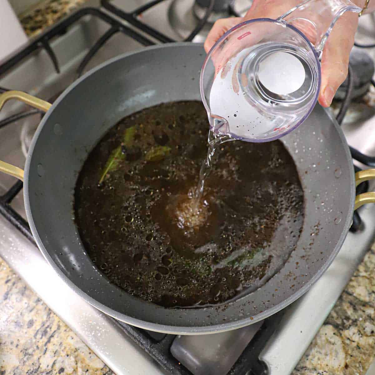 A person pouring water from a measuring cup into a large skillet on a gas stove that is filled with a simmering soy sauce and vinegar mixture.
