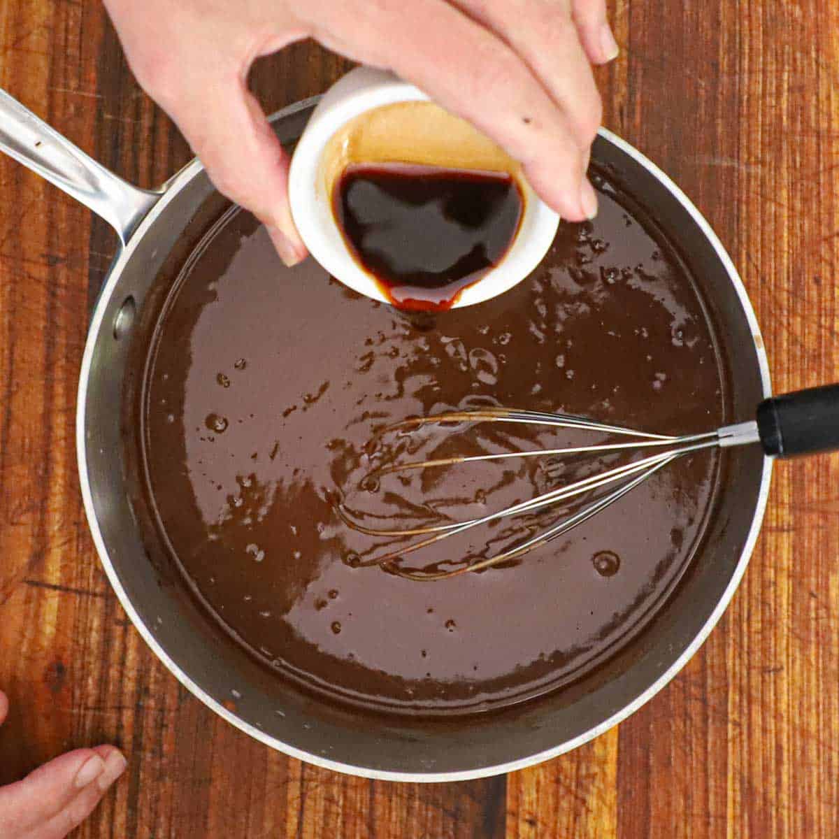 A person pouring vanilla extract from a small white bowl into a saucepan filled with a chocolate custard for homemade chocolate ice cream.
