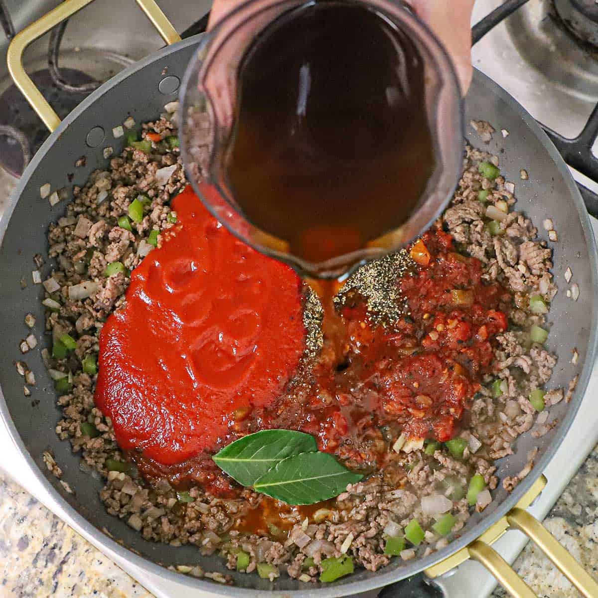A person pouring beef broth from a plastic measuring cup into a skillet that is filled with browned ground beef, sautéed bell peppers and onion, as well as tomato sauce, fire roasted tomatoes, bay leaves, and seasonings.
