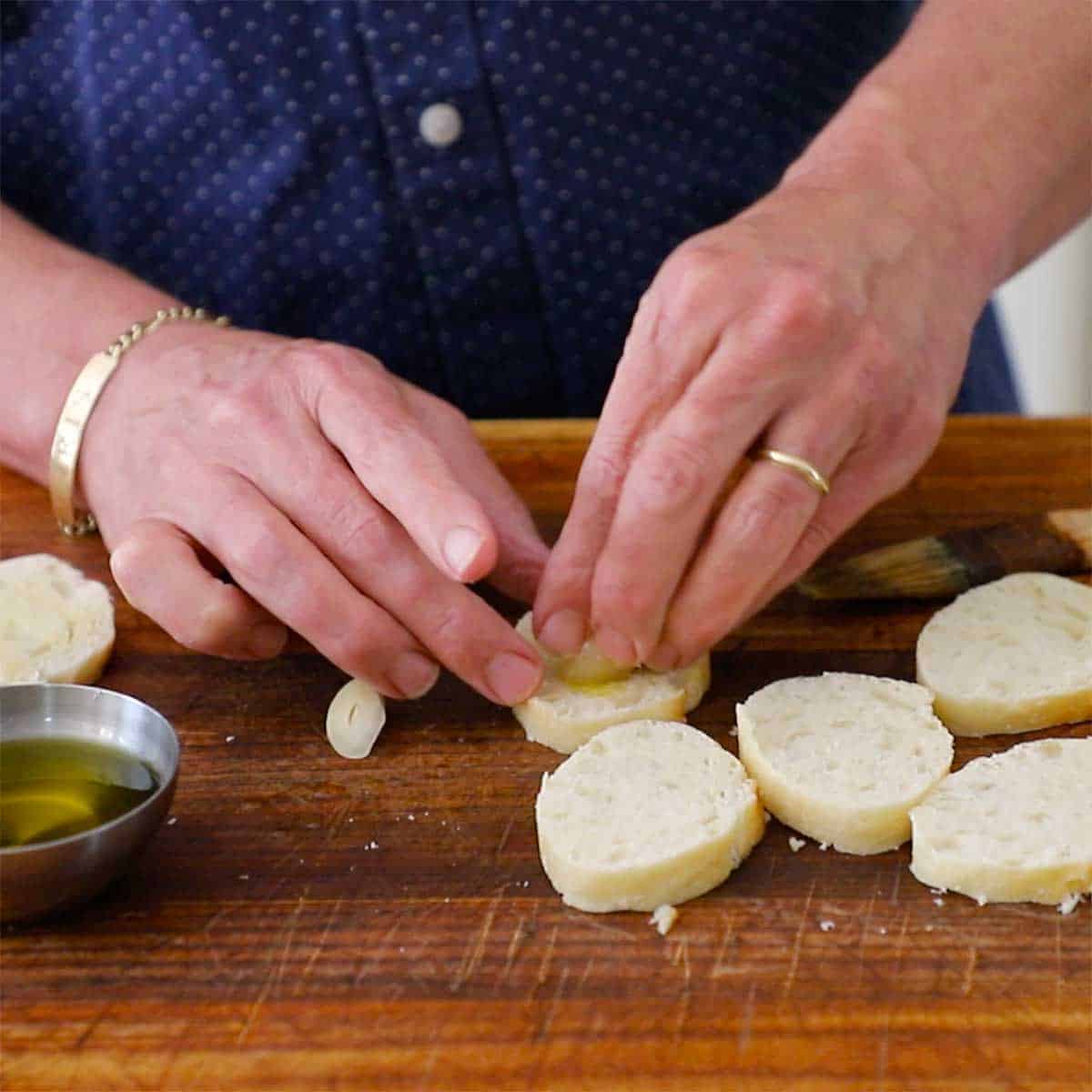 A person rubbing a clove of garlic that has been halved onto the surface of sliced baguette next to other slices that are all resting on a large wooden cutting board.