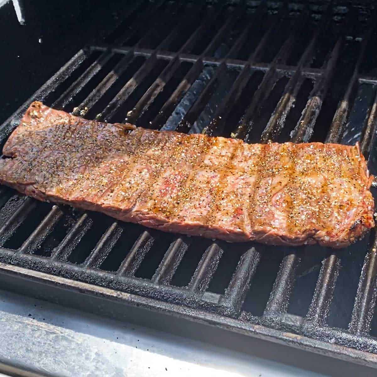 A seasoned skirt steak being grilled over a gas flame on an outdoor gas grill.